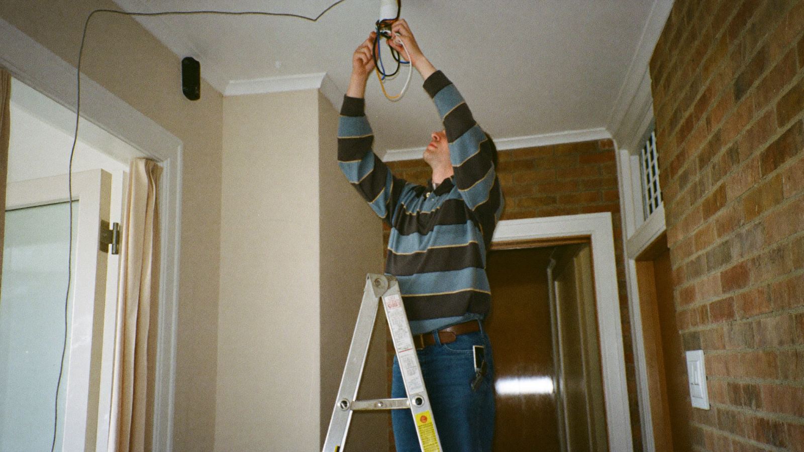 An electrician up a stepladder wiring a ceiling light, phone lighting up in his back pocket