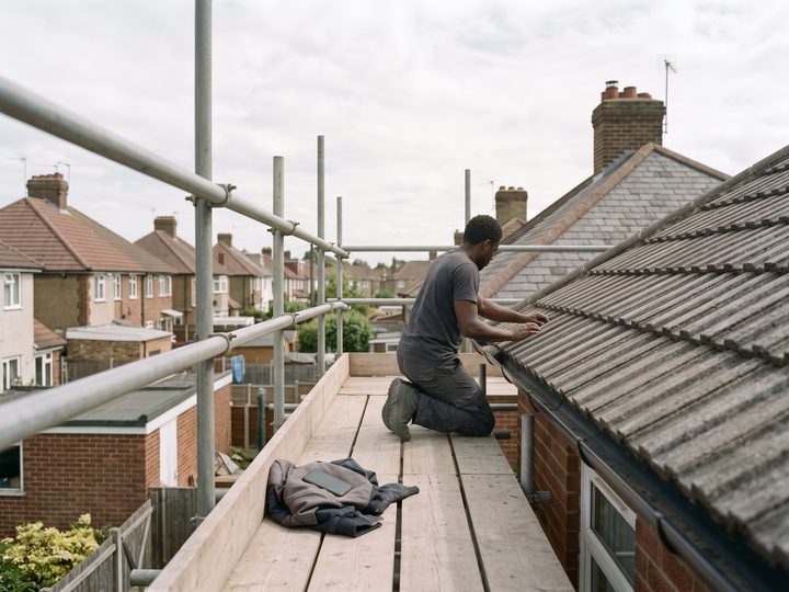 A roofer kneeling on scaffold at the eaves of a house, fixing grey roof tiles